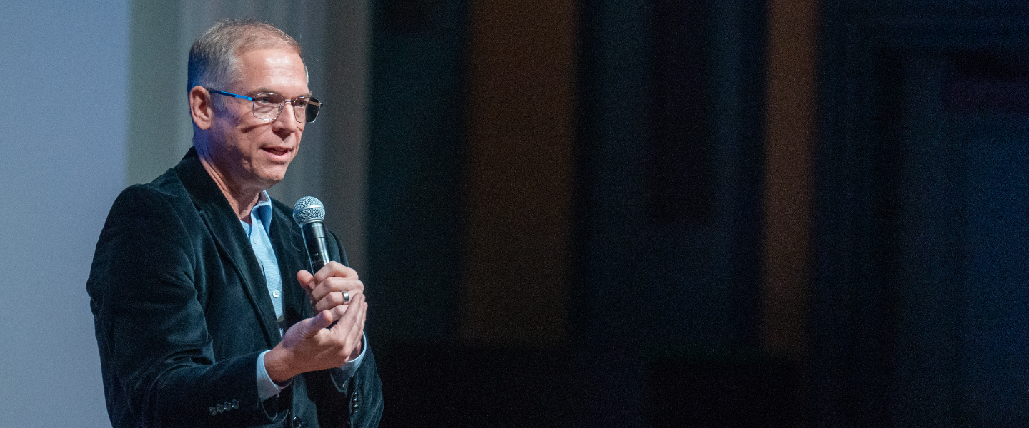 Man in glasses and suit jacket speaking into a microphone, giving a presentation at an event.