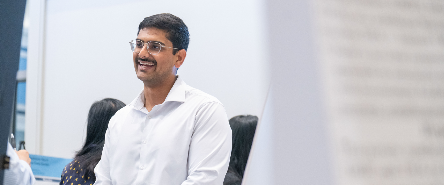 Smiling man with glasses and a white shirt in a professional setting, possibly at a presentation or conference.