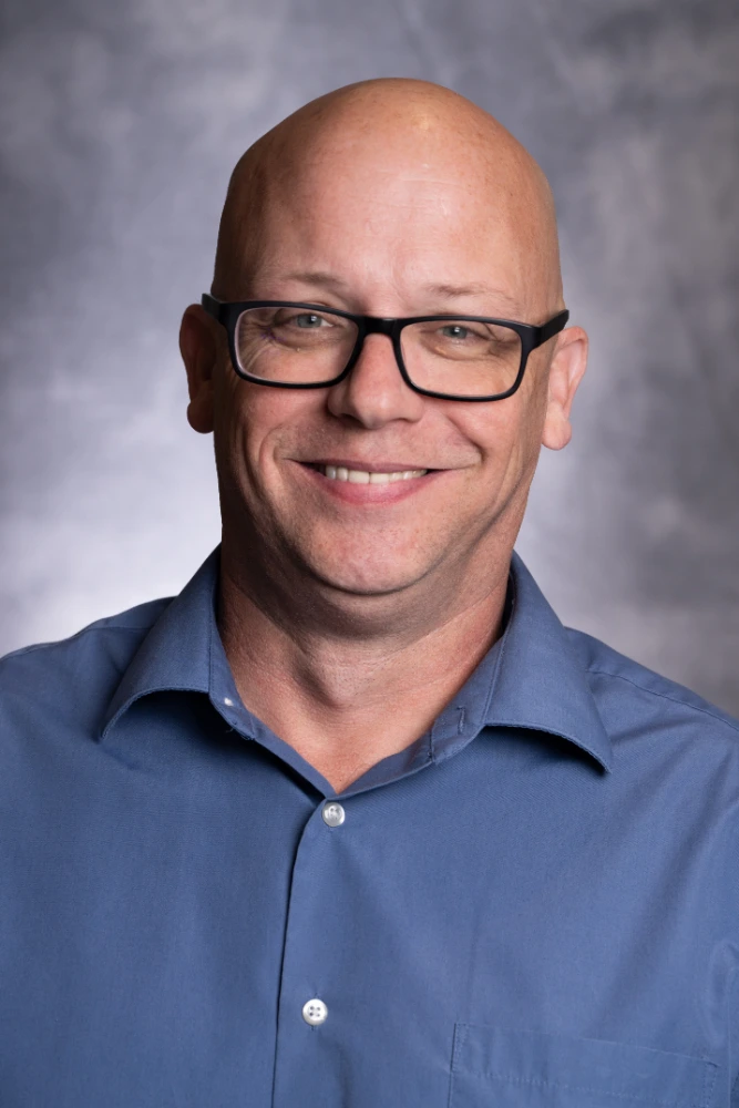 Smiling bald man wearing black rectangular glasses and a blue button-down shirt against a gray studio backdrop.