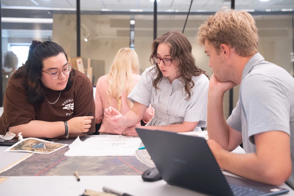 Students and alumni collaborating on a design project, discussing plans at a table with a laptop.