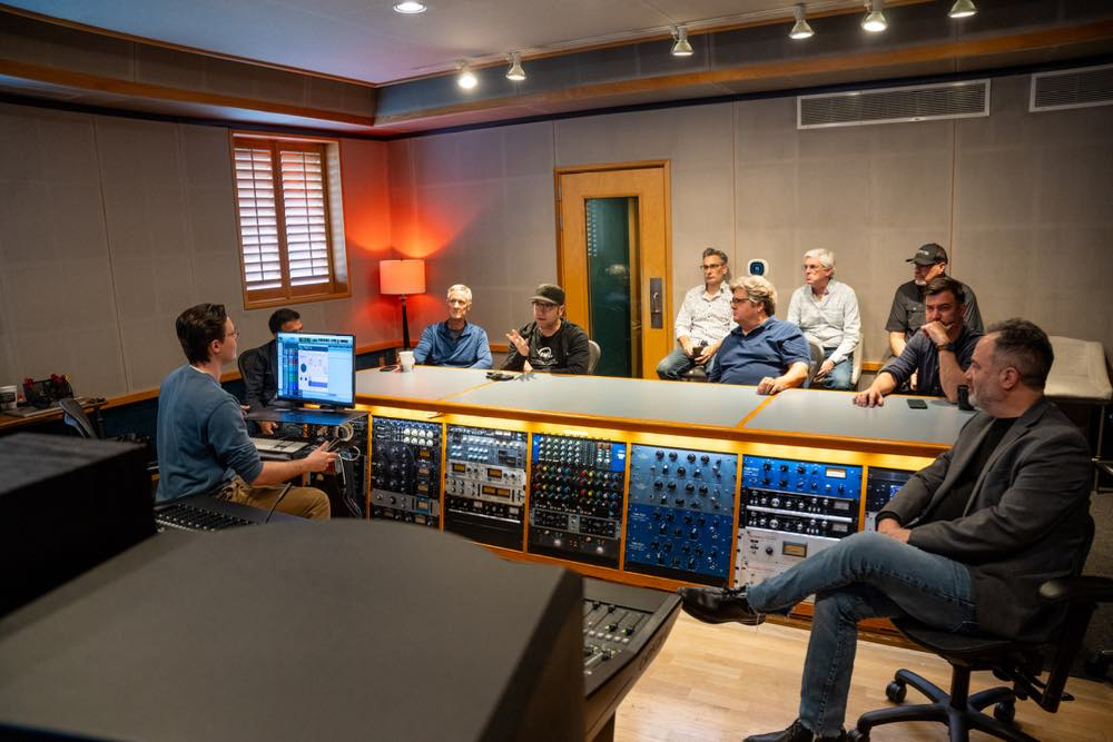 Sound engineer operates a mixing console while a team observes in a professional recording studio.