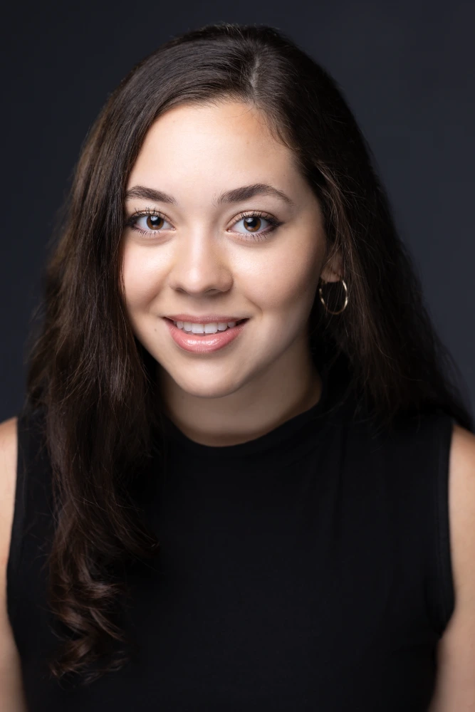 Smiling young woman with long dark hair in a professional headshot, wearing a black top and hoop earrings.