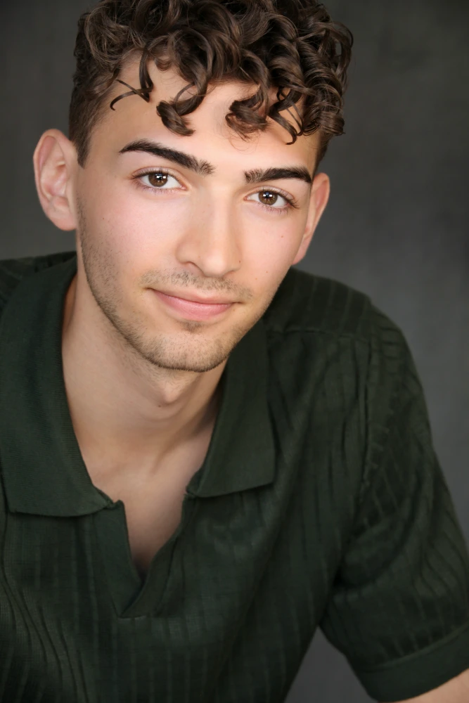 Headshot of a smiling young man with curly brown hair in a dark green shirt against a grey background.