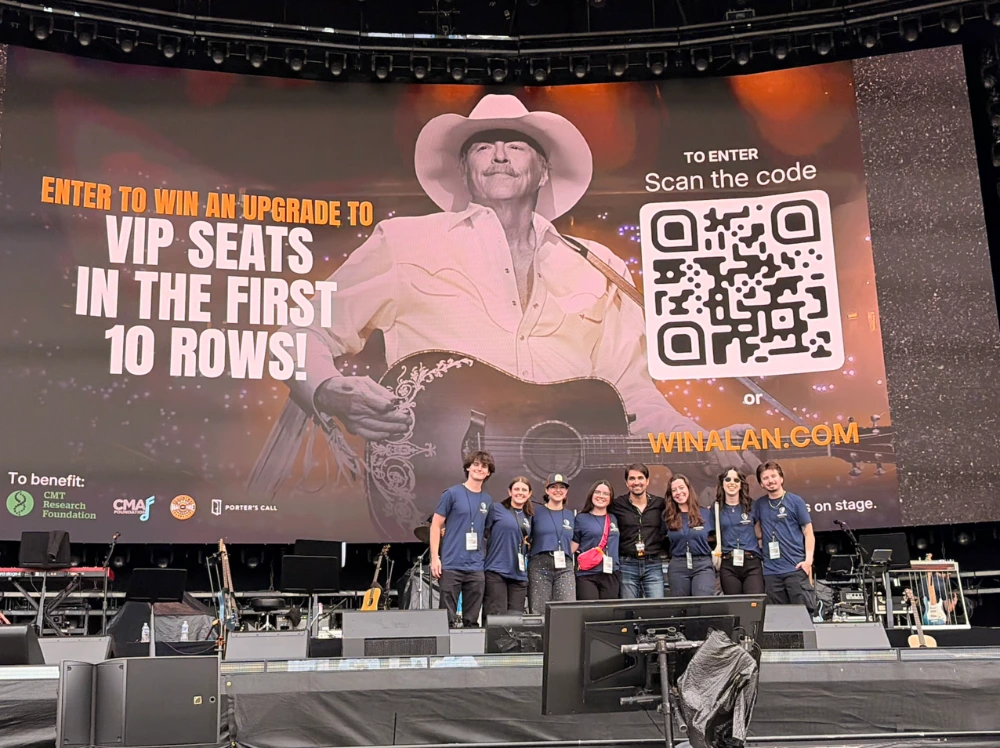 Concert stage with a large screen promoting a VIP seats giveaway with a QR code, while a group of people pose together at the front of the stage.