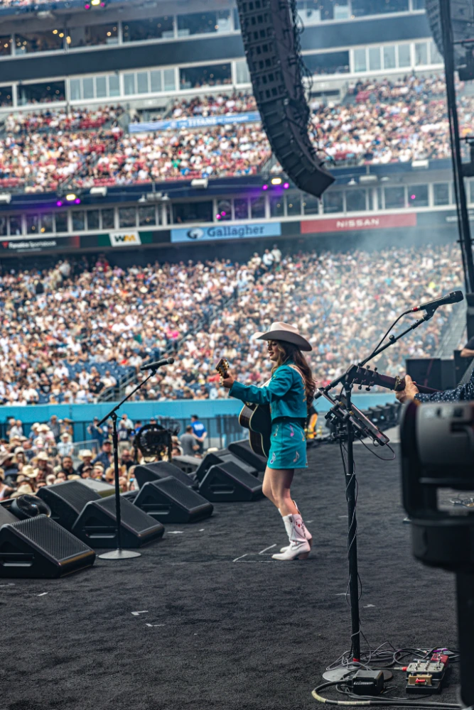 Country singer in a cowboy hat plays guitar on a stadium stage, with microphones and speakers in the foreground and a packed crowd in the stands behind.
