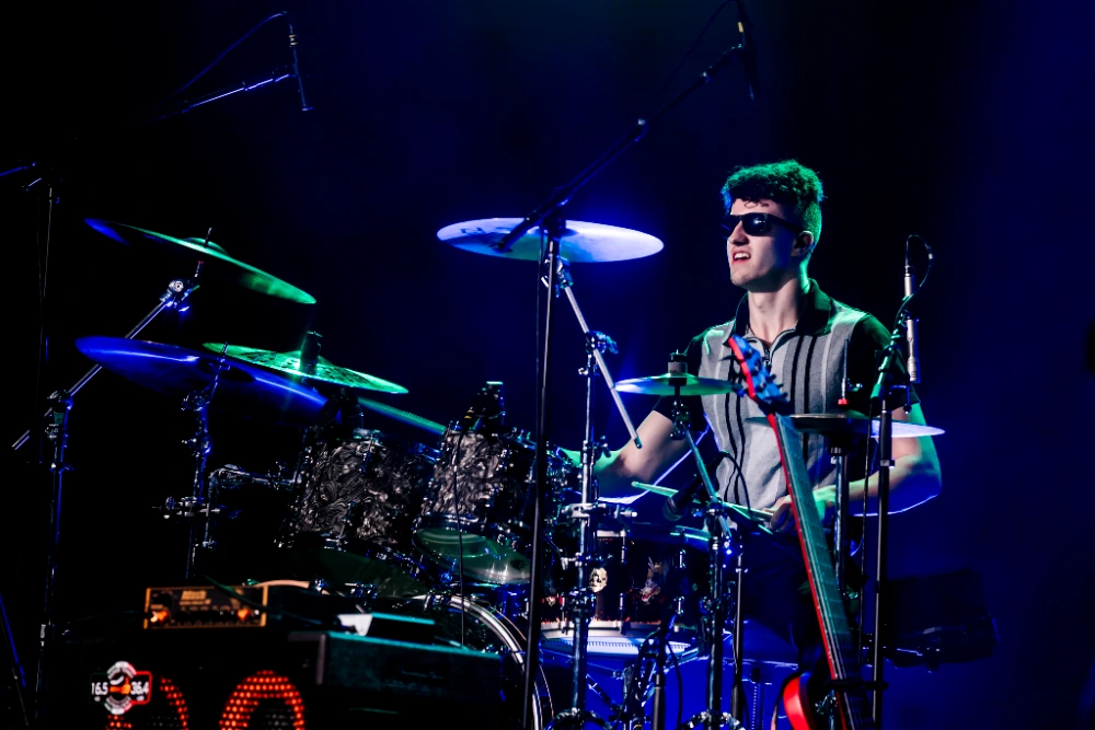 A male drummer wearing sunglasses plays a drum kit on stage under blue and green lights during a live concert.