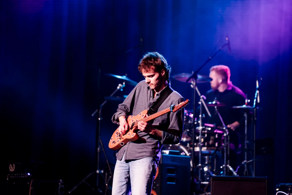 Guitarist playing an electric guitar live on stage, illuminated by blue lights, with a drummer behind.