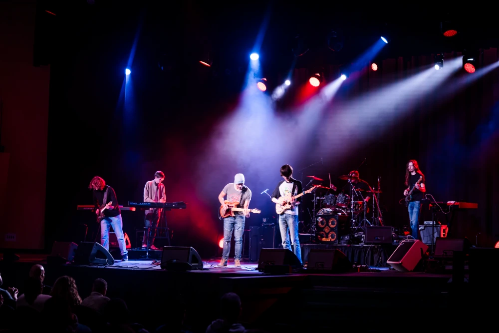 Musicians perform a live concert on stage with vibrant red and blue lighting, playing guitars, keyboard, and drums.