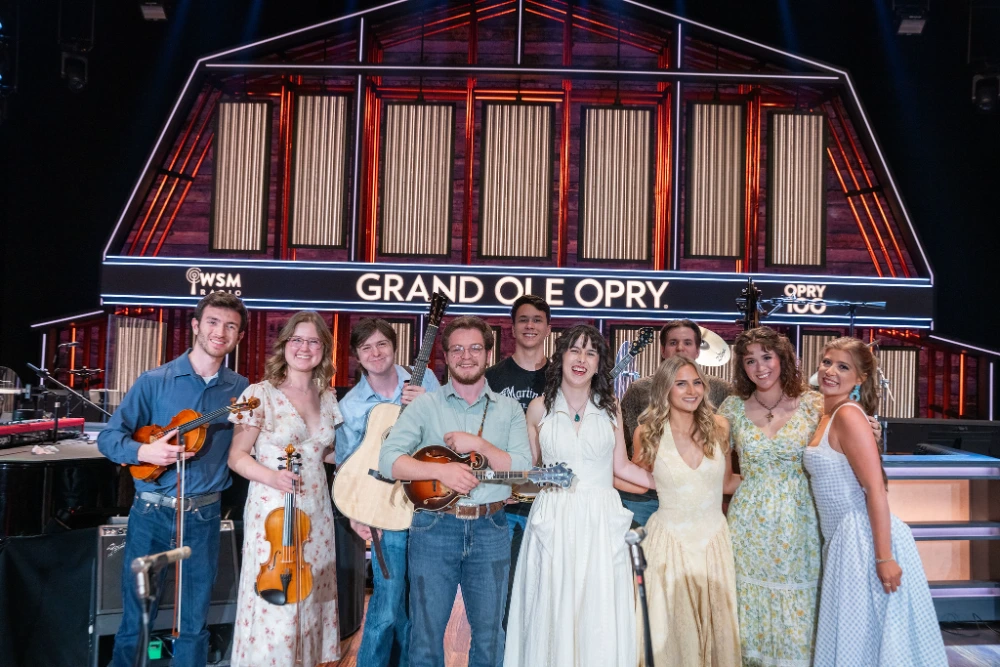 A group of ten young bluegrass and country musicians with fiddles, mandolins, and guitars on the Grand Ole Opry stage.