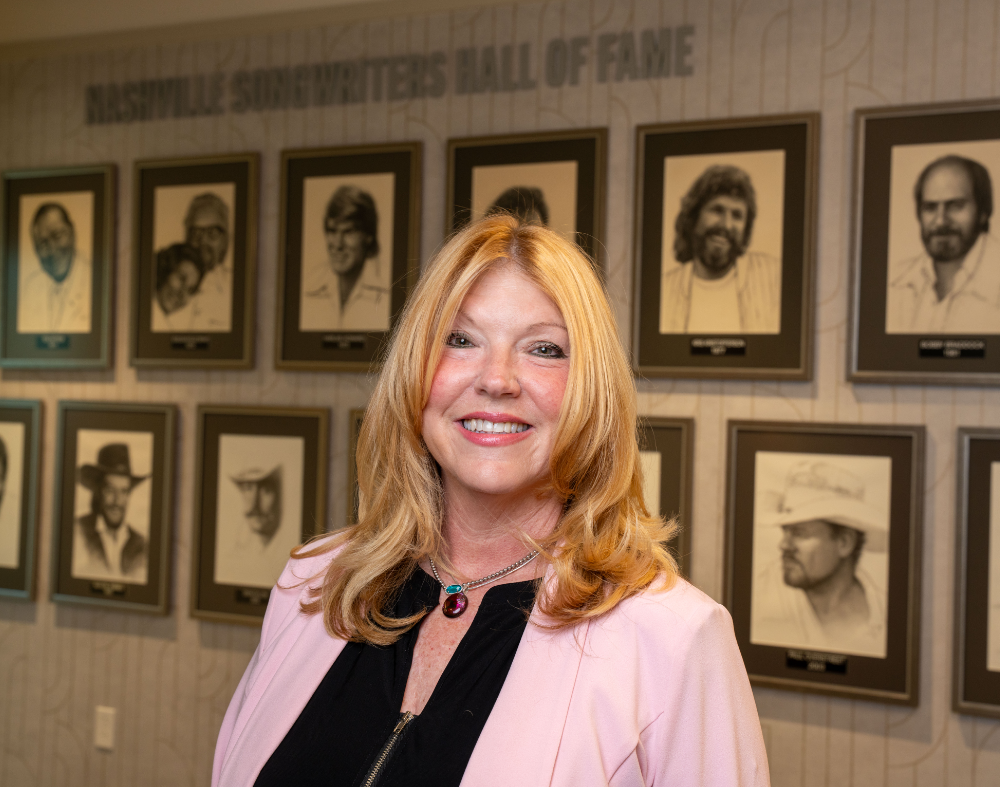 Smiling woman with long blonde hair wearing a pink blazer, against a background of framed portraits.