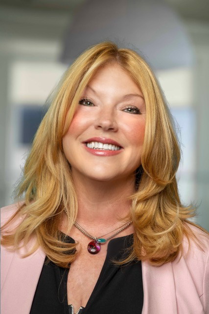 Smiling woman with long blonde hair wearing a pink blazer, against a background of framed portraits.
