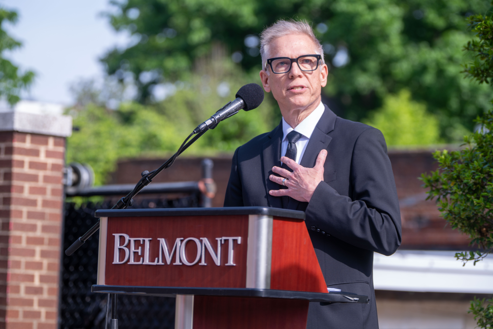 Professional speaker in a suit and glasses at a Belmont University podium, addressing an outdoor event.