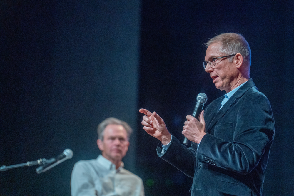 Man in glasses and a dark jacket speaking into a microphone, gesturing, at a public event or conference.