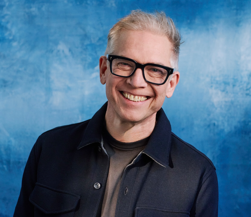 A smiling man with short grey hair and black glasses wears a dark jacket against a blue background.