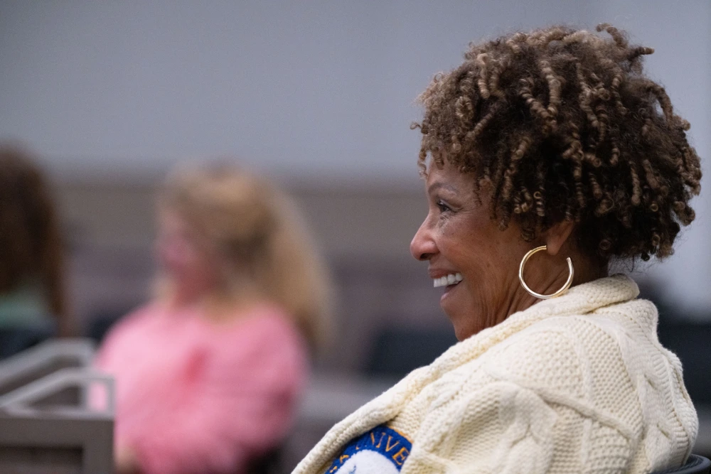 Side-profile of a smiling woman with short curly hair and hoop earrings, seated indoors with other people blurred in the background.