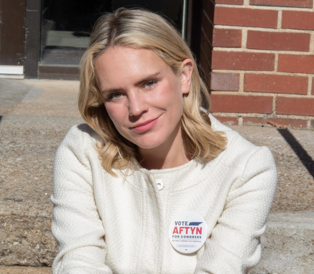 Blonde woman Aftyn Behn, a political candidate, smiling and wearing a 'Vote Aftyn for Congress' button.