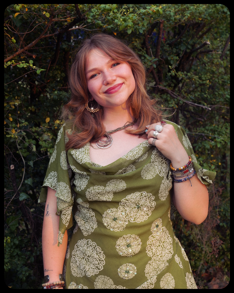 Smiling young woman in a green dress with white floral patterns and bohemian jewelry, standing outdoors.