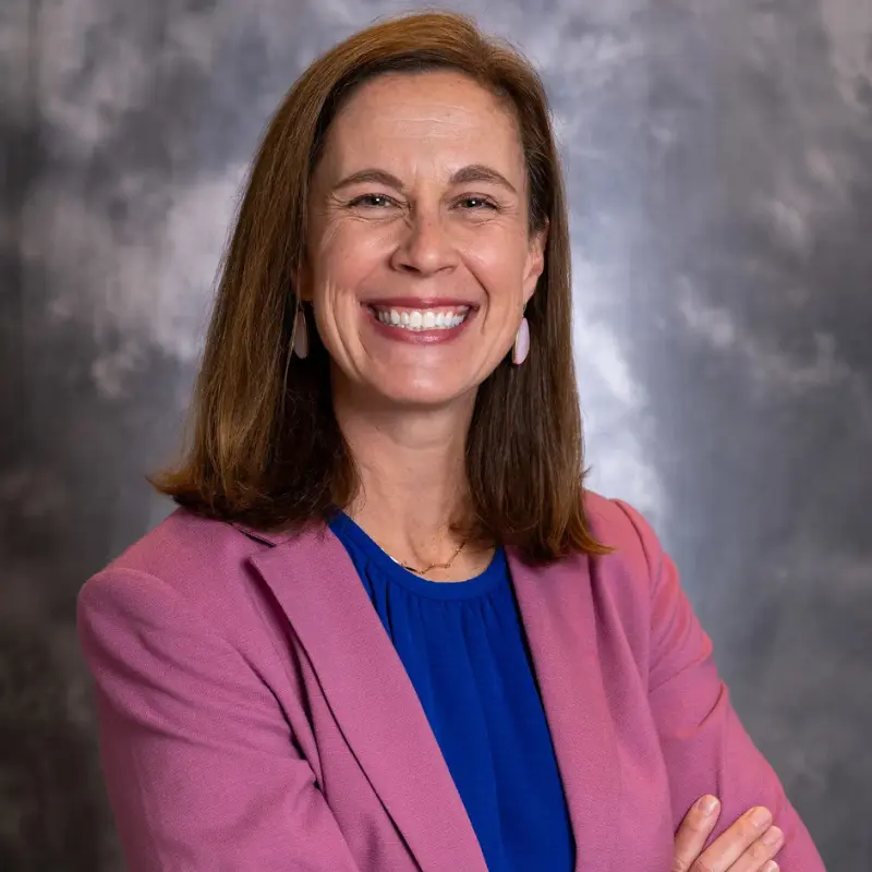 Smiling woman in a pink blazer and blue top, arms crossed, against a gray studio backdrop.