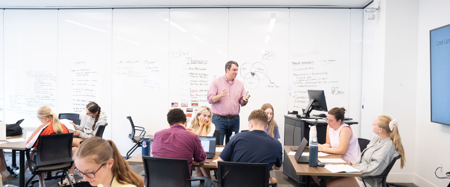 A man in a pink button-up shirt stands and speaks to a classroom of students seated at tables with laptops and notebooks. Behind him, whiteboard walls are covered in handwritten notes and a Venn diagram, and a wall-mounted screen is visible at right.