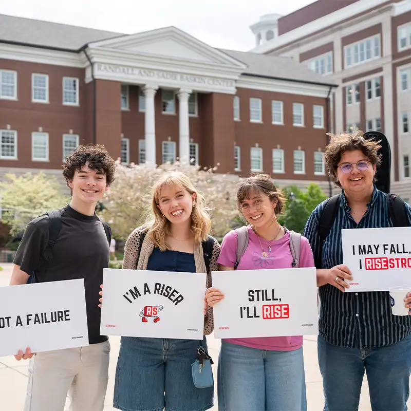 Four college students standing in front of a campus building, each holding a sign that reads (from left to right) “Not a failure,” “I’m a Riser,” “Still, I’ll rise,” and “I may fall, I rise strong.”