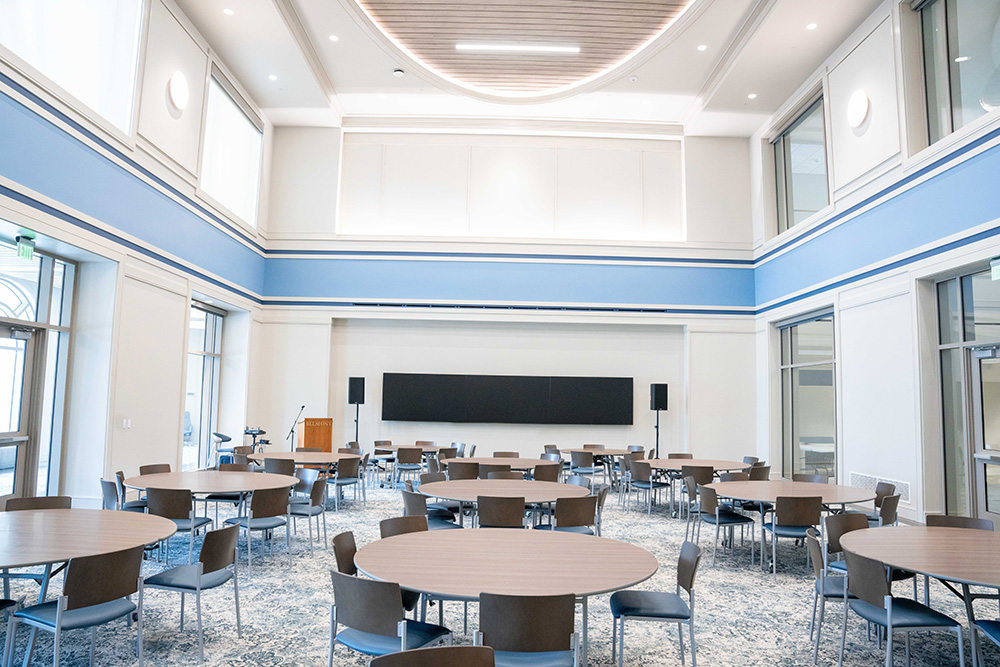 Room filled with round tables, chairs, speakers, a screen, and a podium