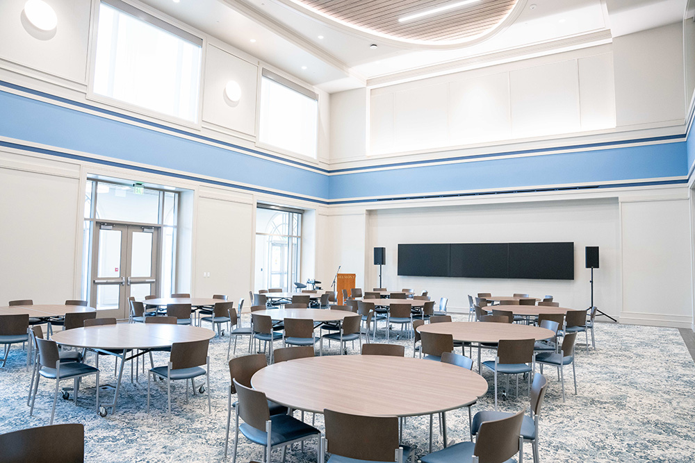 Room filled with round tables, chairs, speakers, a screen, and a podium