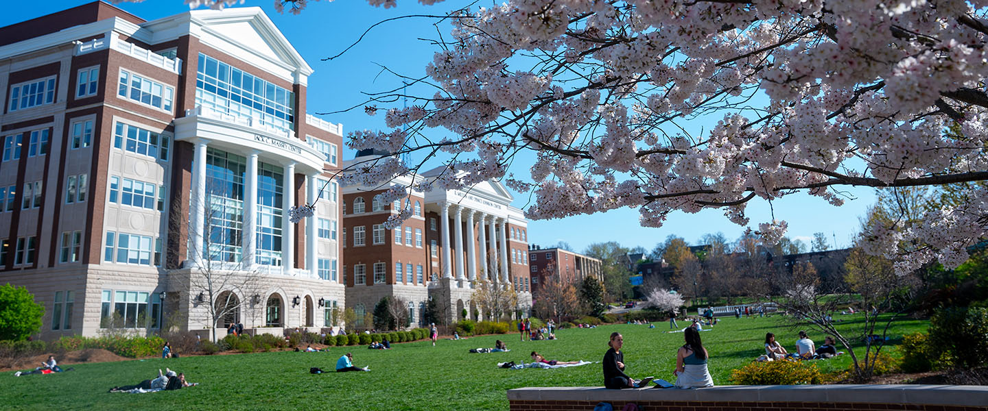 A sunny spring day on campus with students relaxing on the grass in front of Belmont University's Jack C. Massey Center. Cherry blossoms frame the scene.