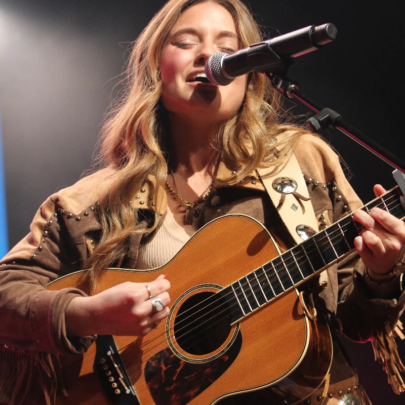Woman with long hair singing into a microphone, playing an acoustic guitar on stage, wearing a brown studded jacket.