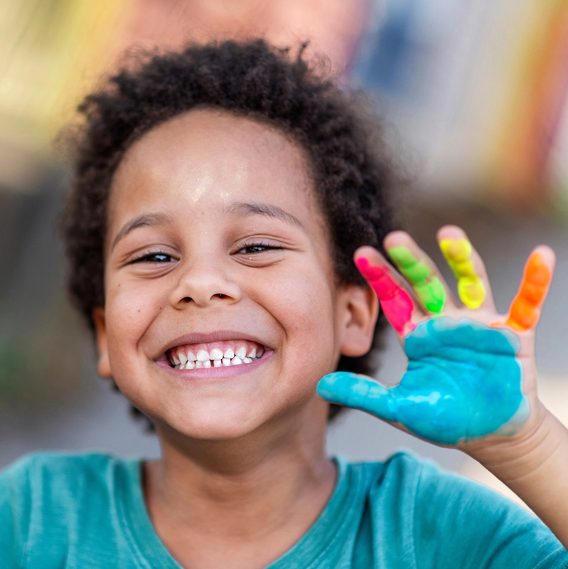 A smiling child with curly hair holds up both hands, covered in bright, colorful paint, showing off the handprints with excitement during a summer camp activity.