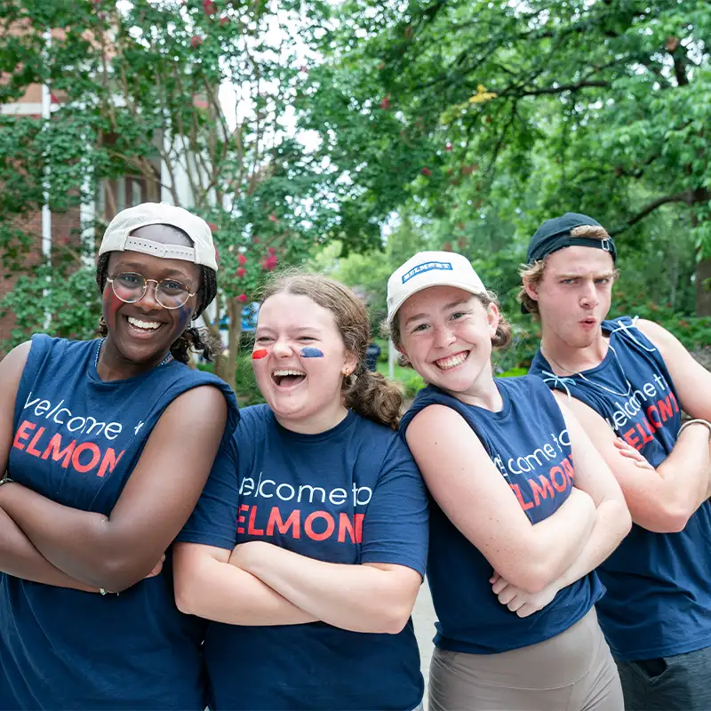 Four smiling young adults stand side by side outdoors with their arms crossed. They’re all wearing navy blue sleeveless T-shirts that say “Welcome to Belmont.” From left to right: a Black woman with glasses and a backward white baseball cap; a white woman with red and blue face paint on her cheeks; a white woman wearing a white cap; and a white man in a black cap making a playful expression. Lush green trees and a brick building are visible behind them.
