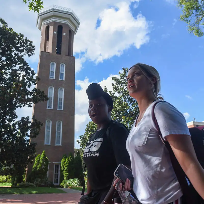 Two young women walking on a pathway in front of the Belmont Bell Tower, a tall brick structure. The woman on the left has a bun hairstyle and wears a black shirt, while the woman on the right is in a white shirt and holds a phone. The sky is partly cloudy, with trees and greenery surrounding the area.
