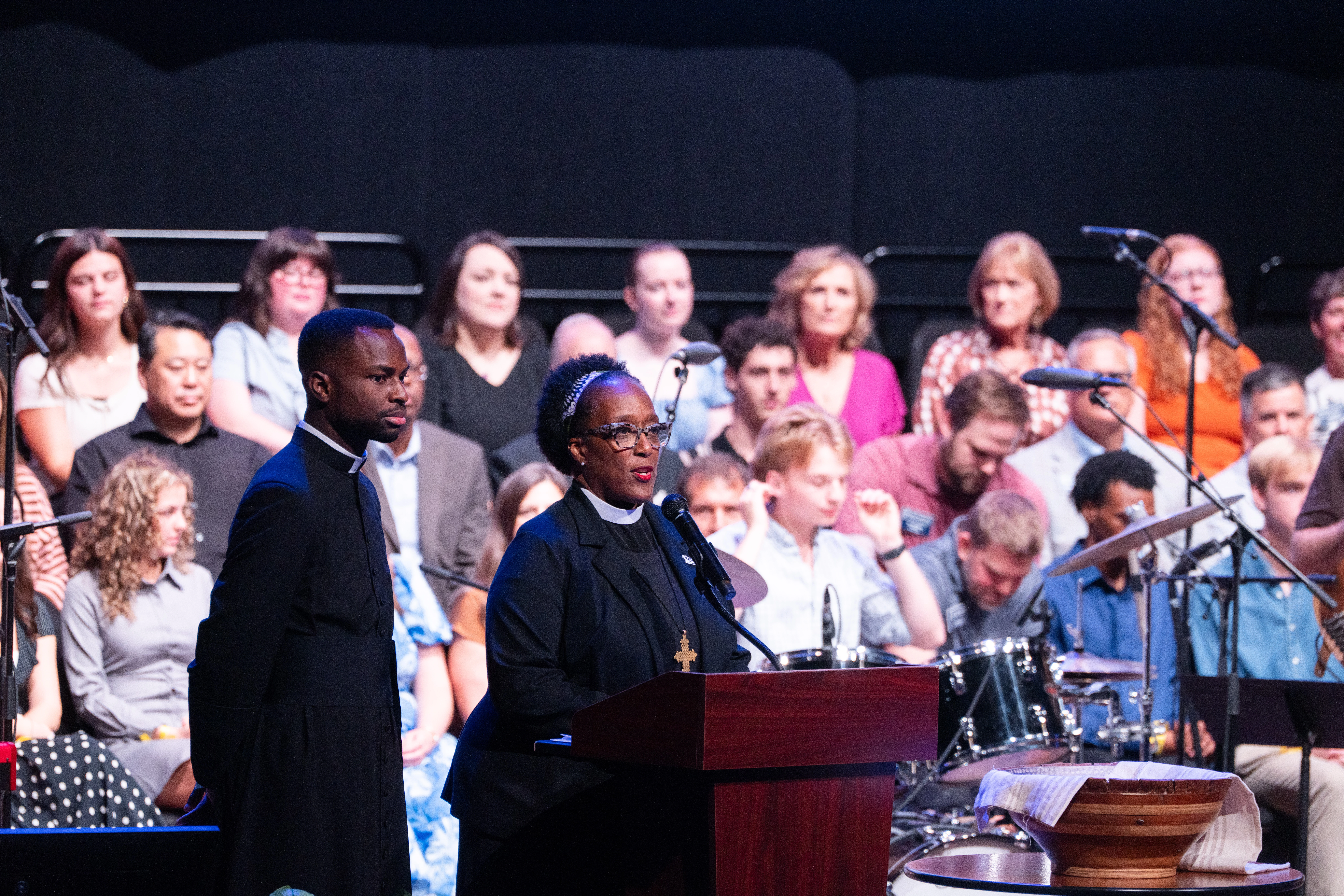 A woman and man stand behind podium addressing audience with choir seated behind them