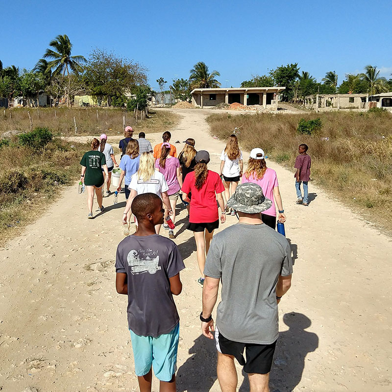 A group of students walk with children along a dirt road in a village setting.