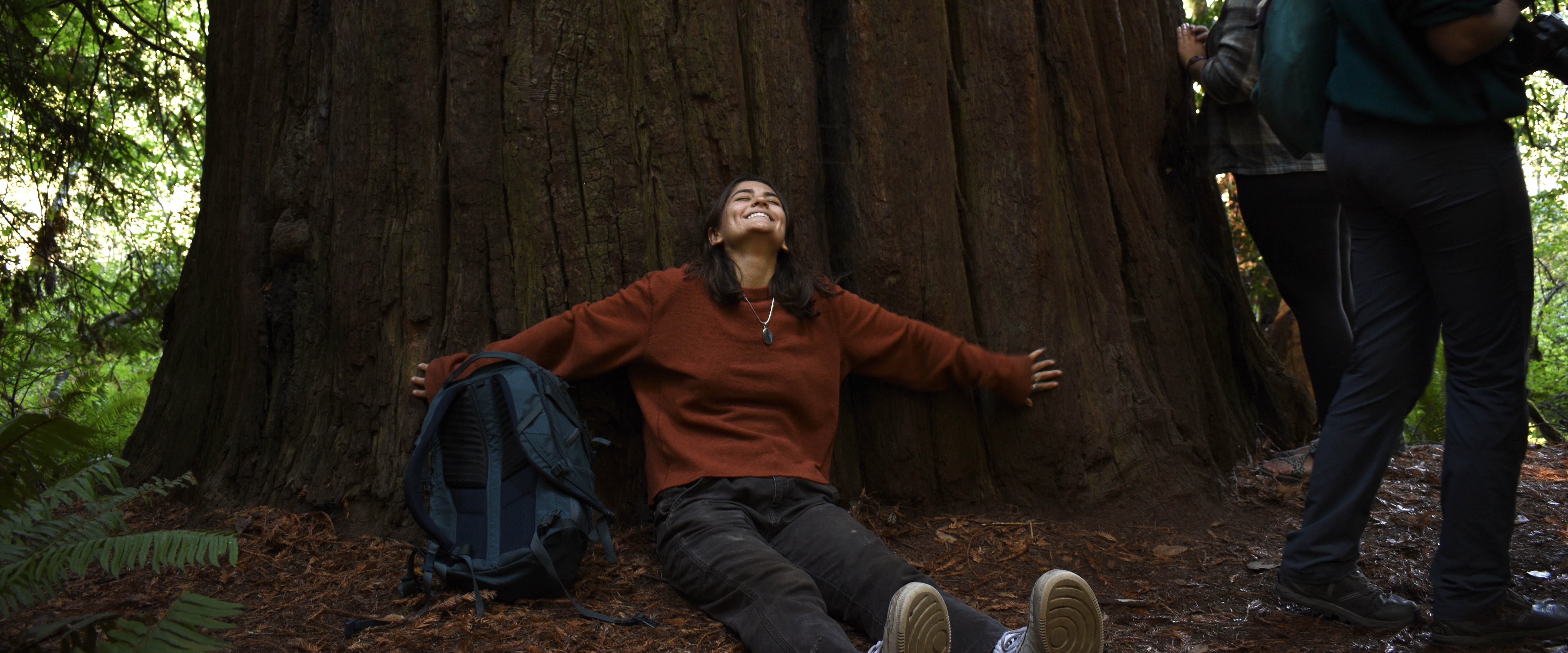 Belmont USA student sits against a large Red wood tree with her arms out and head looking up to the sky