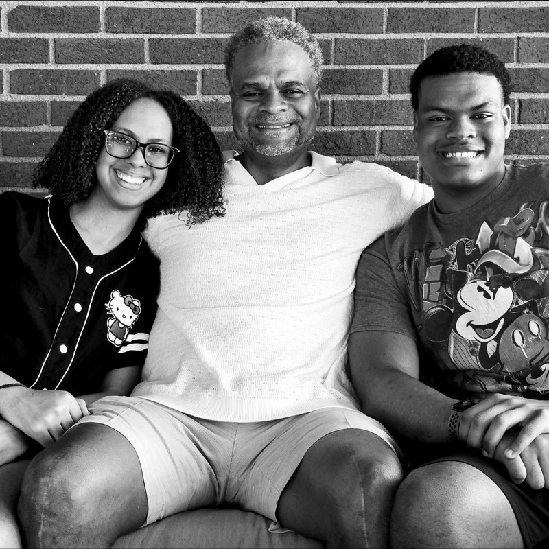 A black-and-white photo of three people sitting close together and smiling in front of a brick wall.