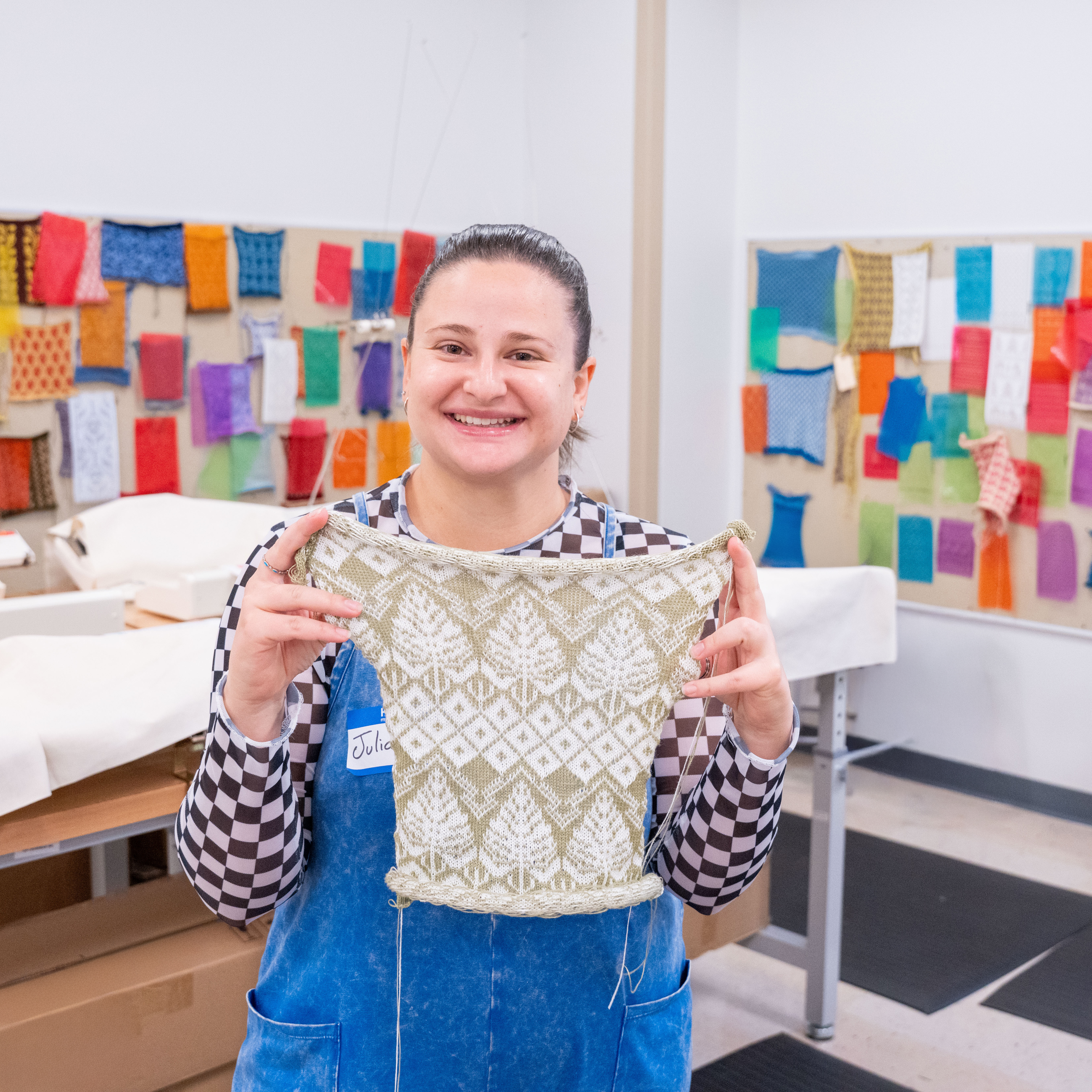 Woman with brown hair holding up a knitted pieces of cloth in front of a board of other cloth samples. 