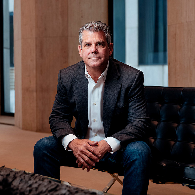 Middle-aged man in a blazer and white shirt sitting on a black leather chair in a modern office setting, looking at the camera.