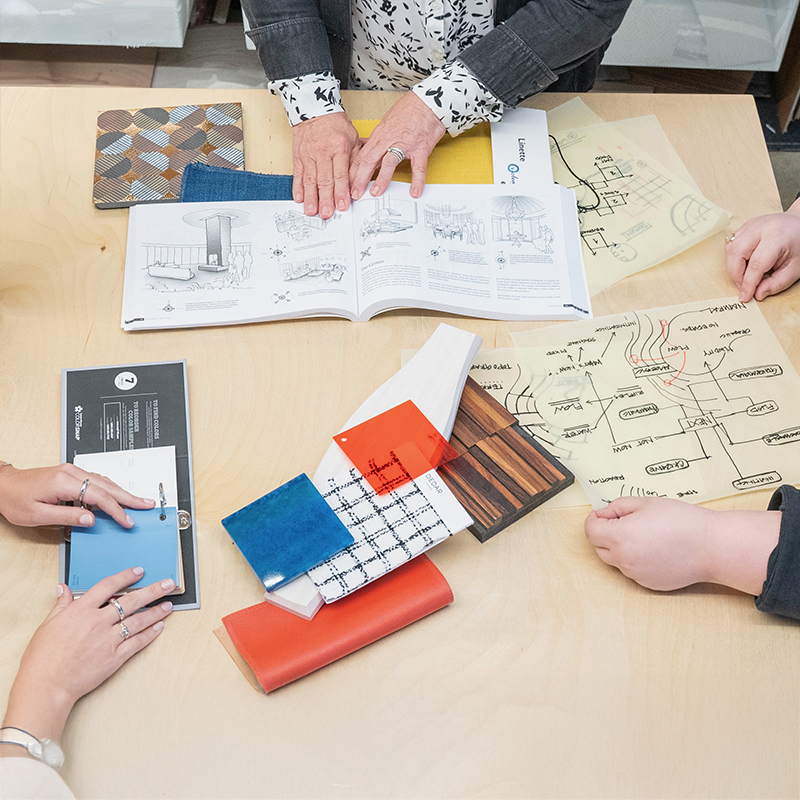 Texture samples for interior design on a table top. The image is cropped and shows three people standing around the table, only their hands are shown. 