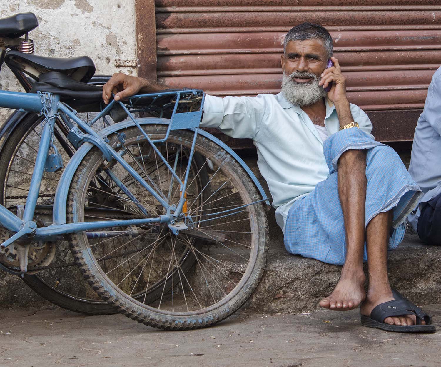 Indian man sitting on the ground next to a blue bike while he talks on his phone